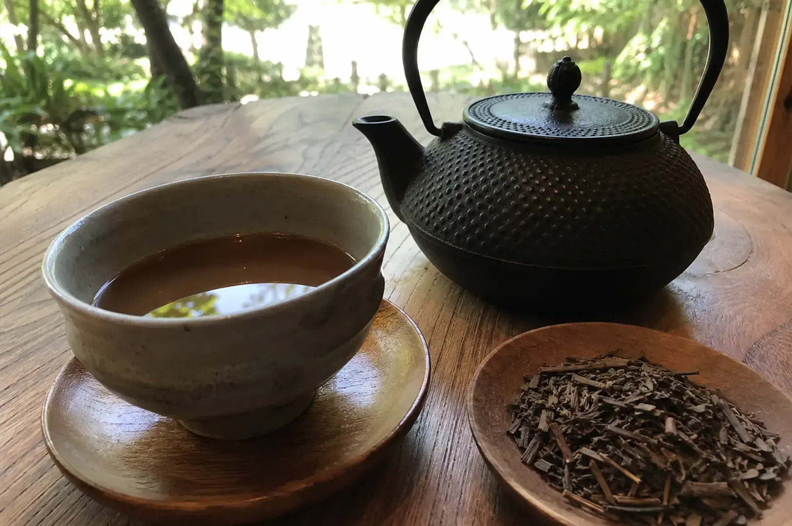 A black, textured iron teapot is paired with a matching cup of tea and a small wooden plate filled with loose, roasted tea stalks, all arranged on a wooden table overlooking a blurred garden view.