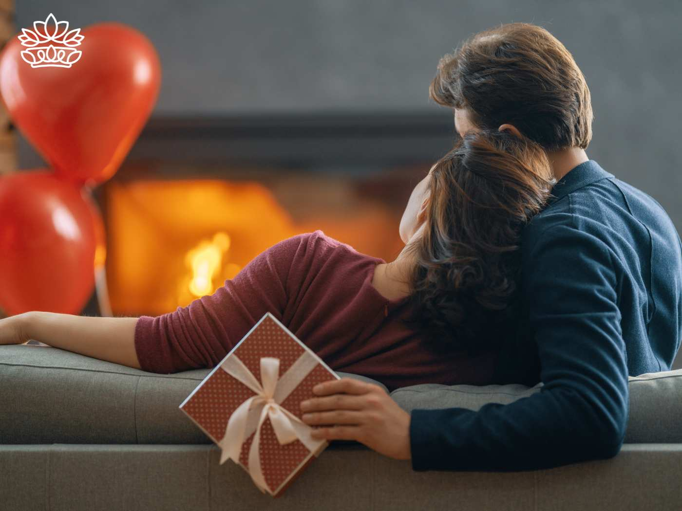 Couple embracing in a cosy living room with a romantic fireplace and Valentine's Day balloons in the background, holding a gift that symbolises love and care. Fabulous Flowers and Gifts. Delivered with Heart.