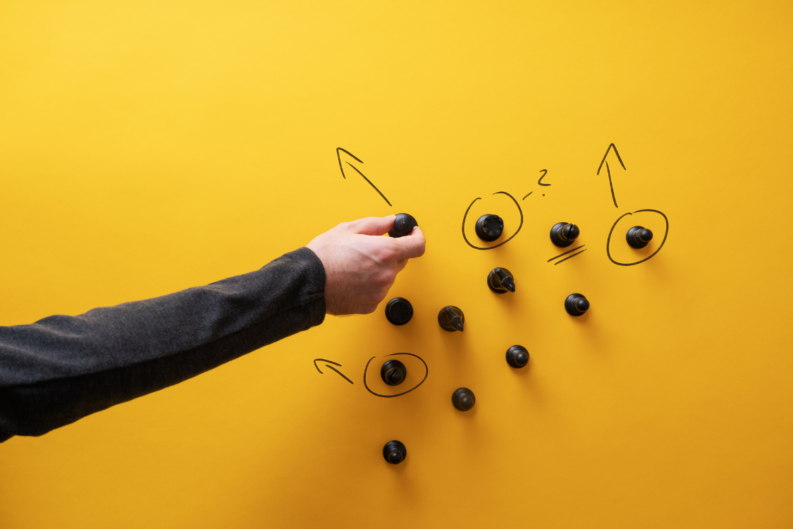 Hand planning a business strategy using chess pieces on a yellow background with arrows and circles drawn around them