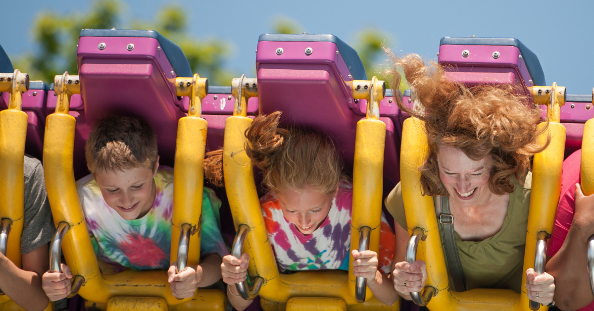 A mom and her two kids laugh and scream with excitement on a roller coaster ride at a Seaside Heights boardwalk amusement park on a bright summer day