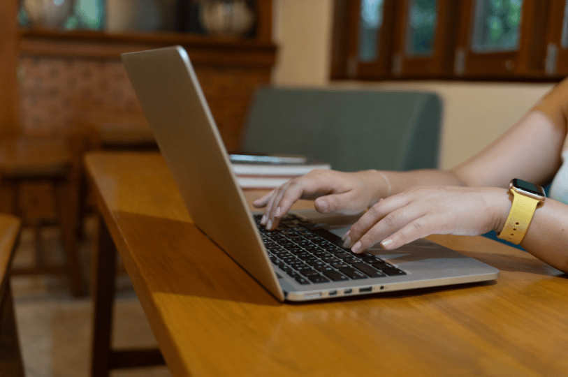 An over-the-shoulder shot of a person working on a laptop at a wooden table in a cafe-like setting, representing the research or implementation phase of an SEO strategy.