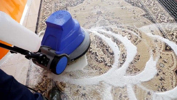 A rotating foam carpet cleaning machine with a blue and gray body applying thick white foam to an ornate beige and brown Persian-style rug.