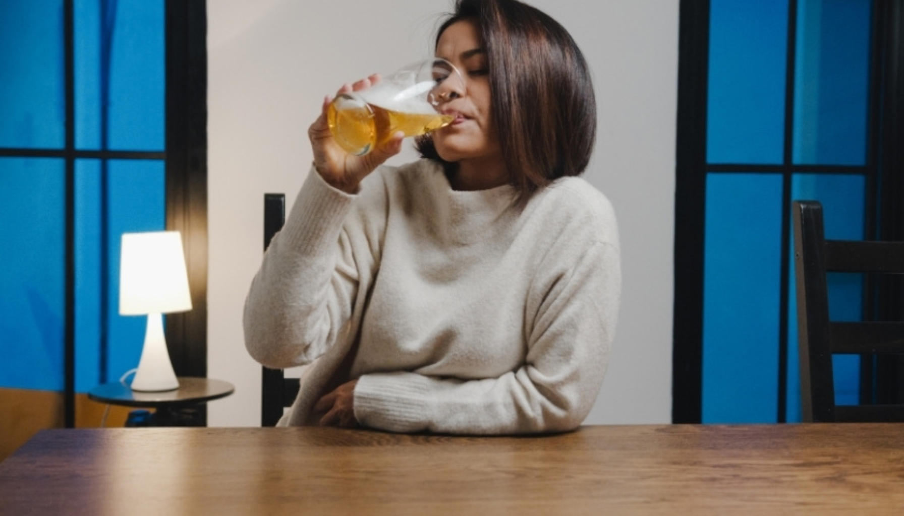 Kvinne i lys genser sitter ved bordet og drikker øl fra et stort glass i et rom med dempet belysning og blå vinduer. Ask ChatGPT