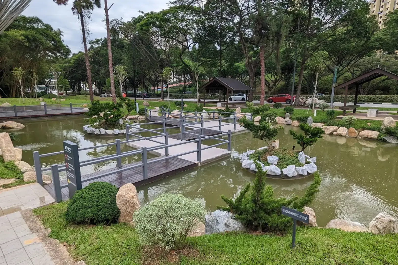 A high-angle view looking down at a zig-zagging grey wooden boardwalk crossing a pond, surrounded by landscaped rock borders, small green island mounds with miniature trees, wooden shelters, and tall trees lining the background.