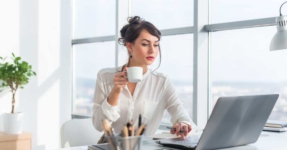 A focused woman holding a cup of coffee while working on her laptop, researching the OnlyFans SETC tax credit.