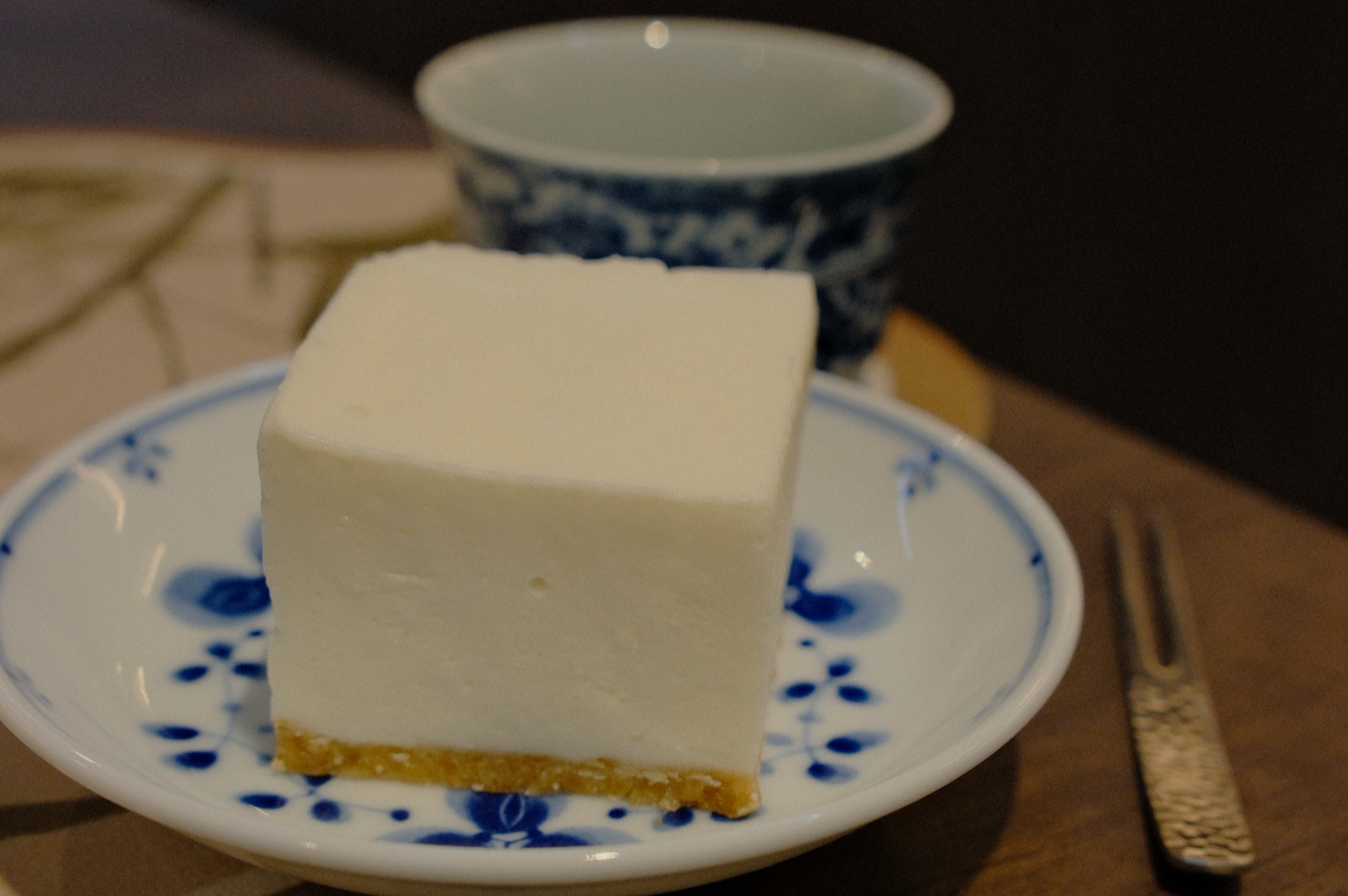 A square slice of cheesecake with a graham cracker crust on a floral-patterned plate, accompanied by a blue-patterned teacup and a fork. Cozy and inviting.