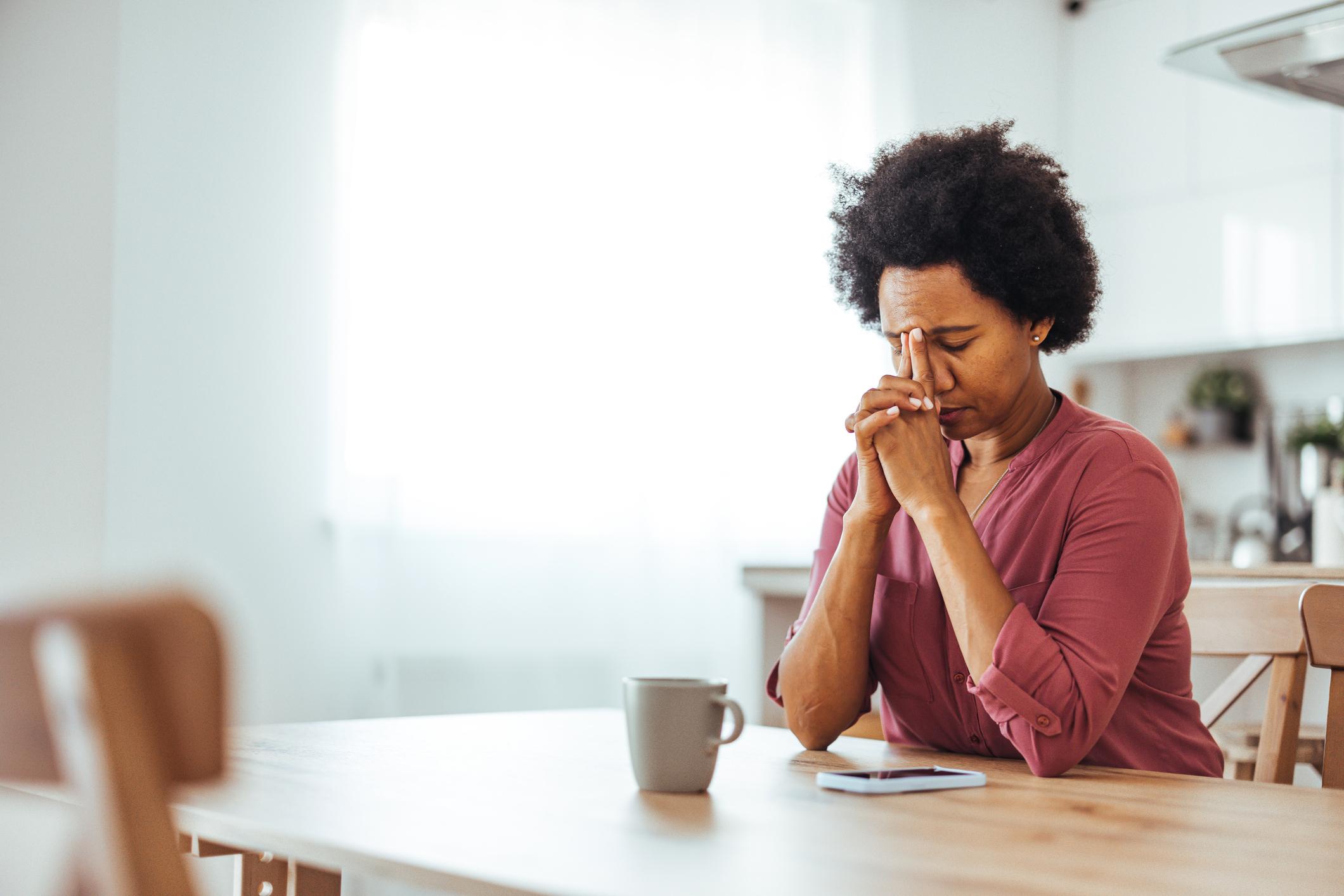 An African-American woman is in a bright kitchen, sitting at her kitchen table, in a visibly uncomfortable posture likely caused by uterine fibroids.