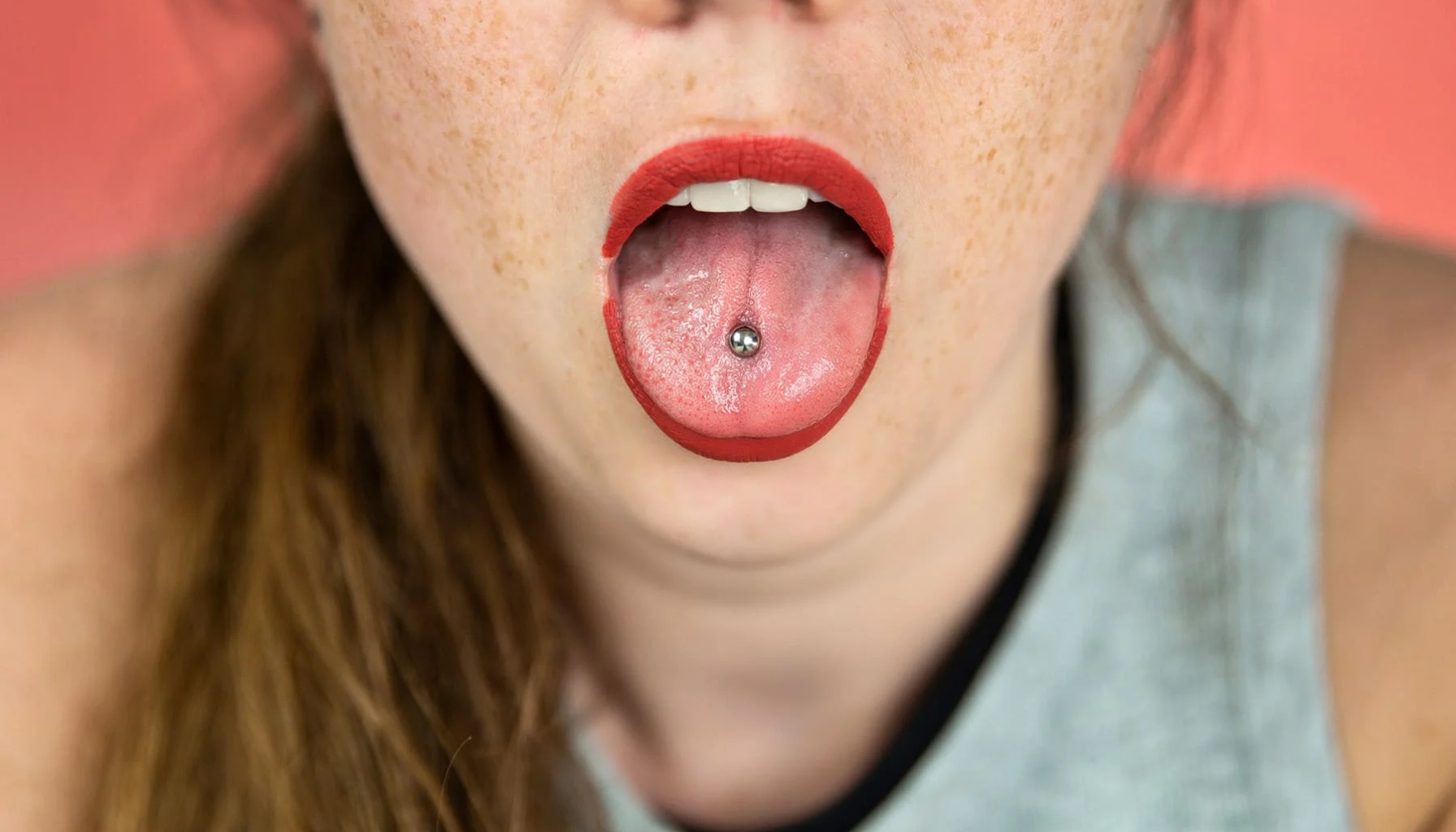 Person med tungepiercing viser frem smykket på tungen med åpen munn, rød leppestift og nøytral bakgrunn.