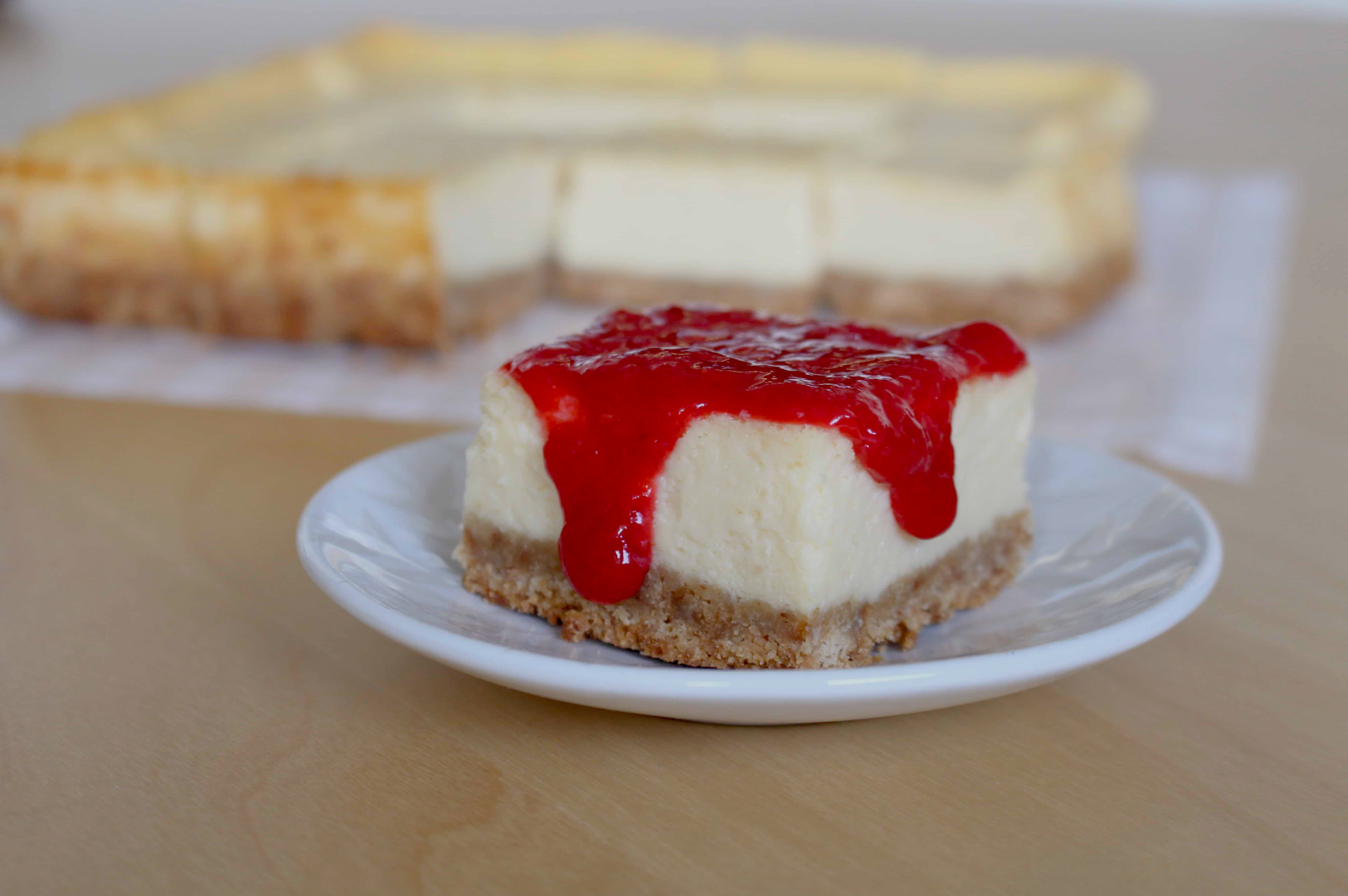 A slice of creamy cheesecake topped with vibrant red strawberry sauce on a plate, with the whole cheesecake blurred in the background, on a wooden table.