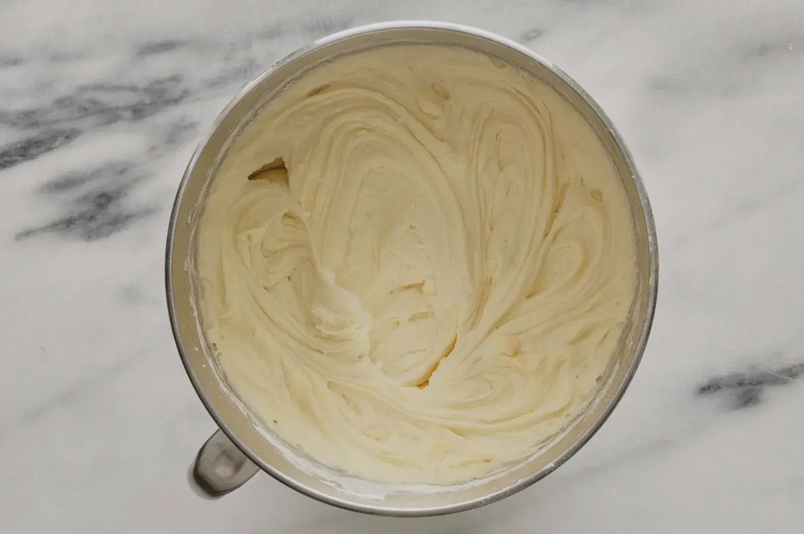 A metal bowl filled with smooth, creamy buttercream frosting, showing swirls and a light beige color.