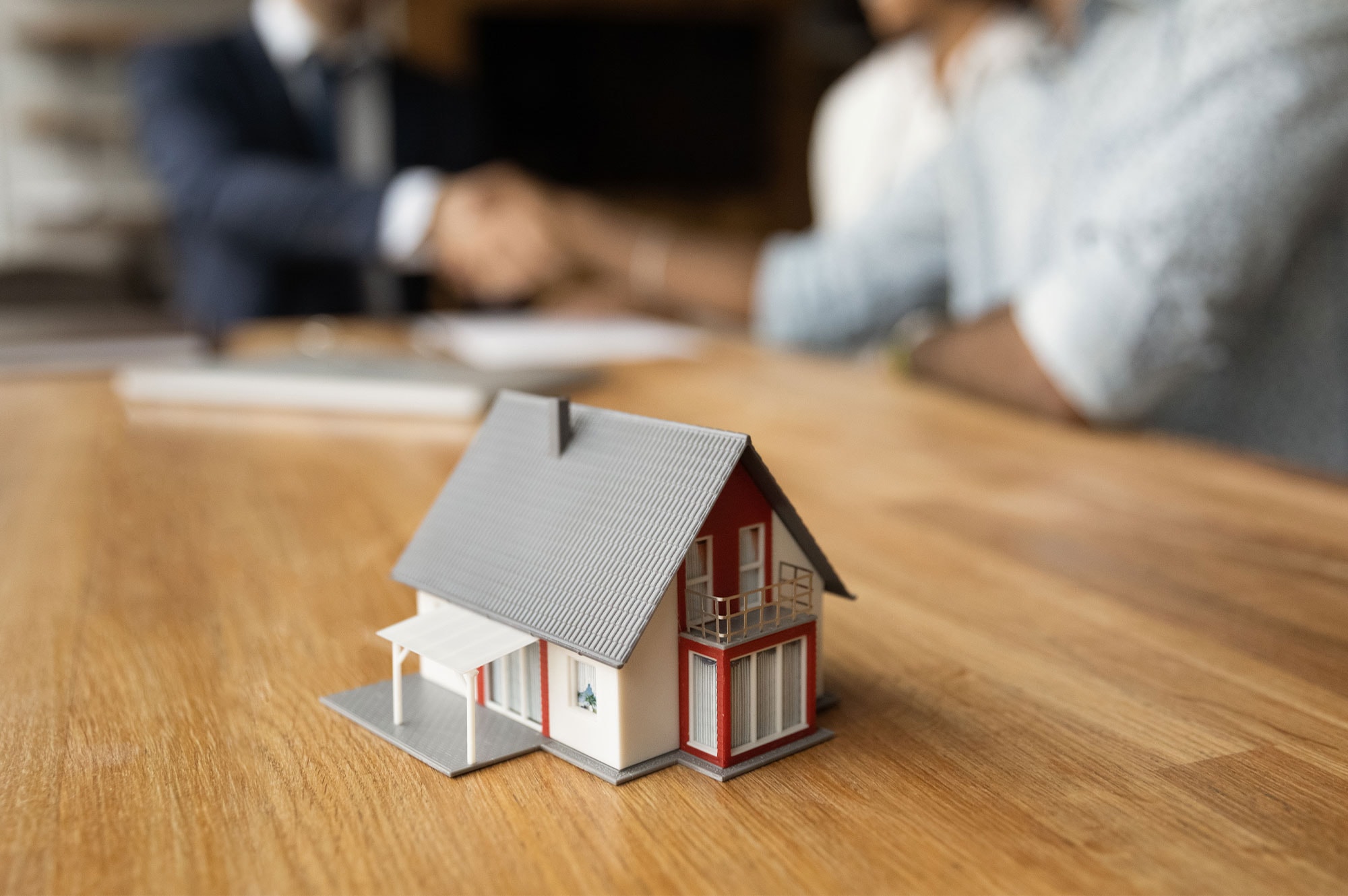 Miniature house on a table with people shaking hands in the background, indicating property purchase agreement