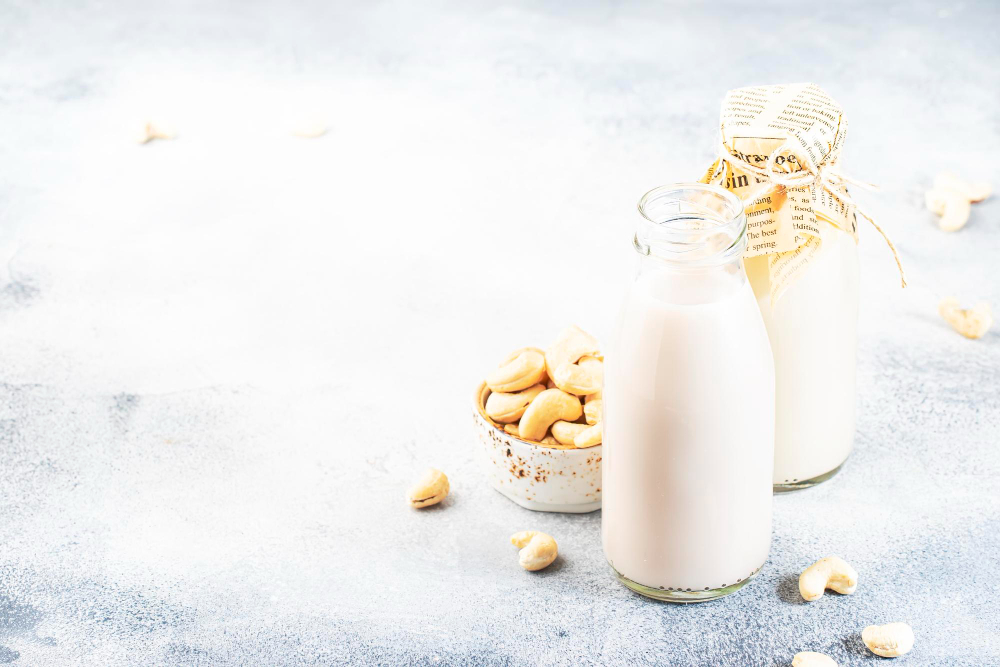 An open and a covered glass bottle of cashew milk with a few cashew nuts near them. 