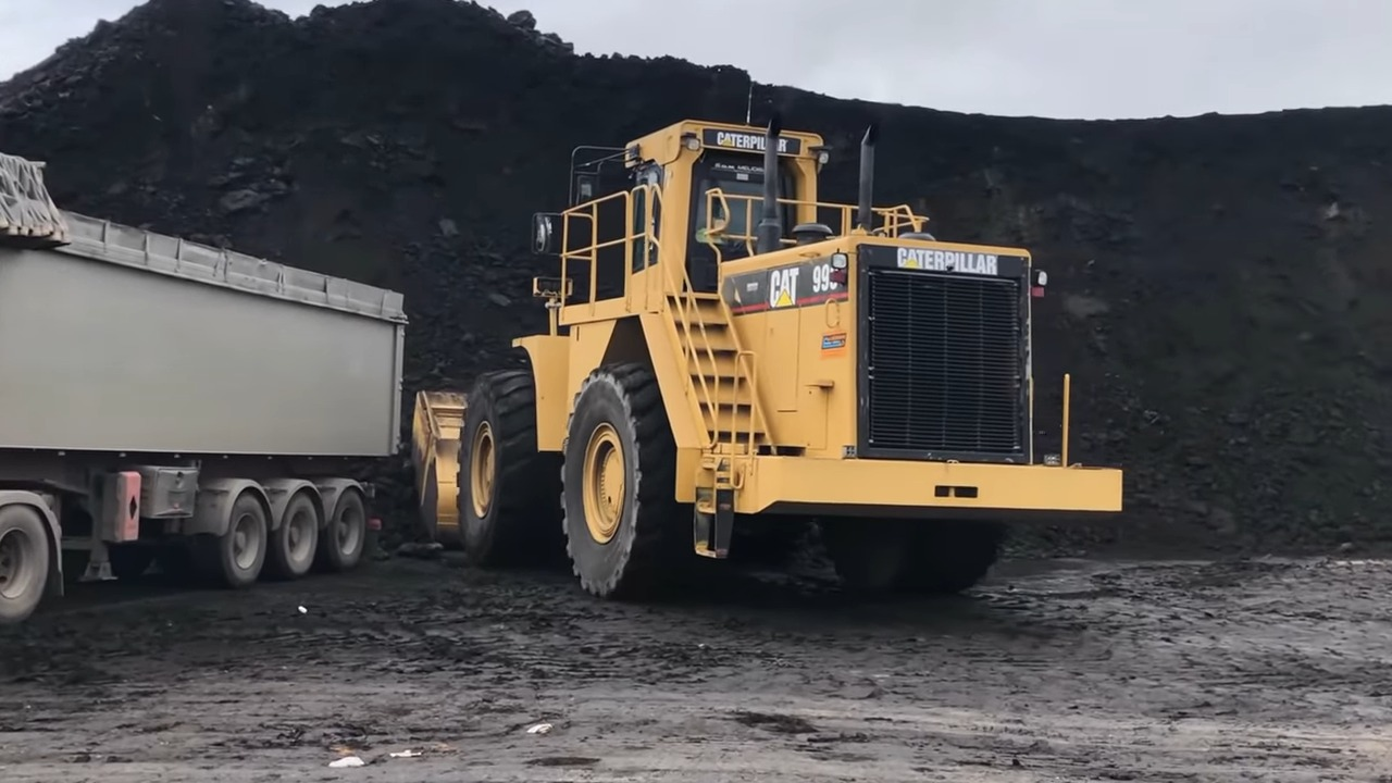 A large yellow Caterpillar front-end loader with the number 994 on its side, parked next to a pile of coal and a truck with a trailer