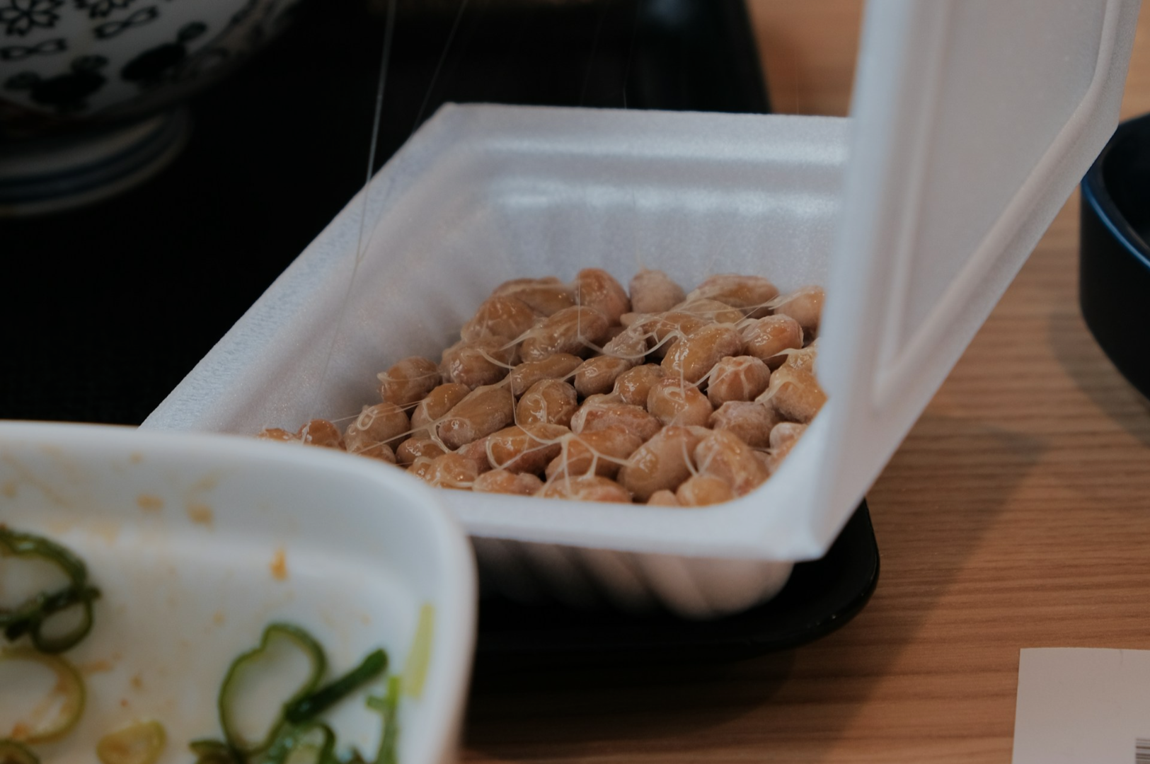 Open styrofoam container of natto with sticky strands, placed on a table. Partially visible bowl of sliced green onions nearby, evoking a simple meal setting.