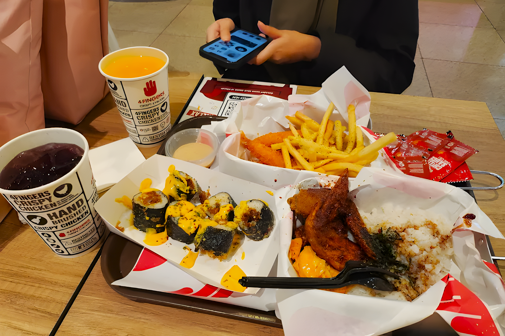 A fast-food meal with sushi, fries, fried chicken, and rice on a tray. Two drinks and sauce packets are nearby. A person is using a phone.