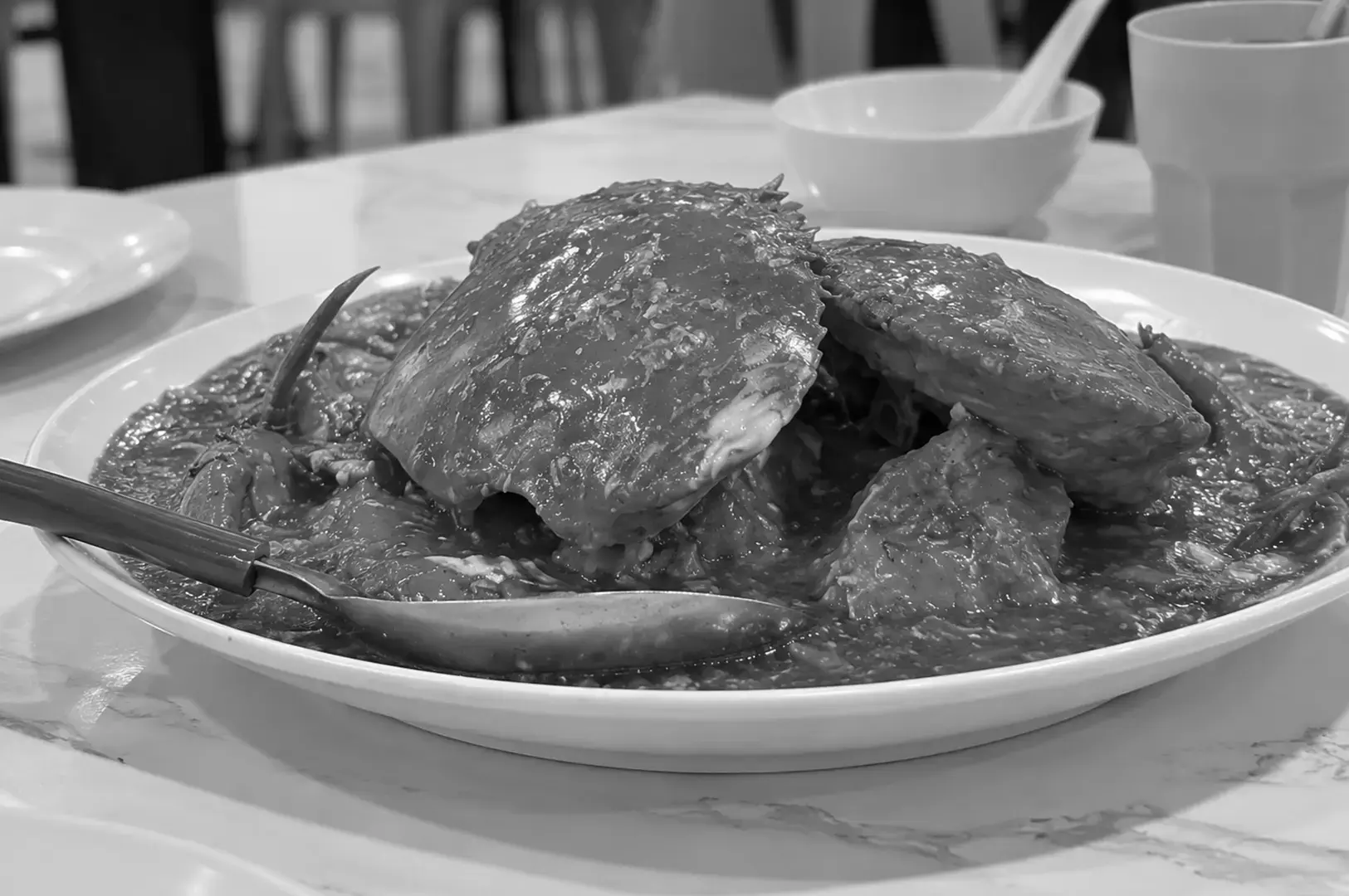 Close-up of a whole chili crab coated in glossy, rich sauce on a white oval plate, with a metal serving spoon placed in front of the dish.