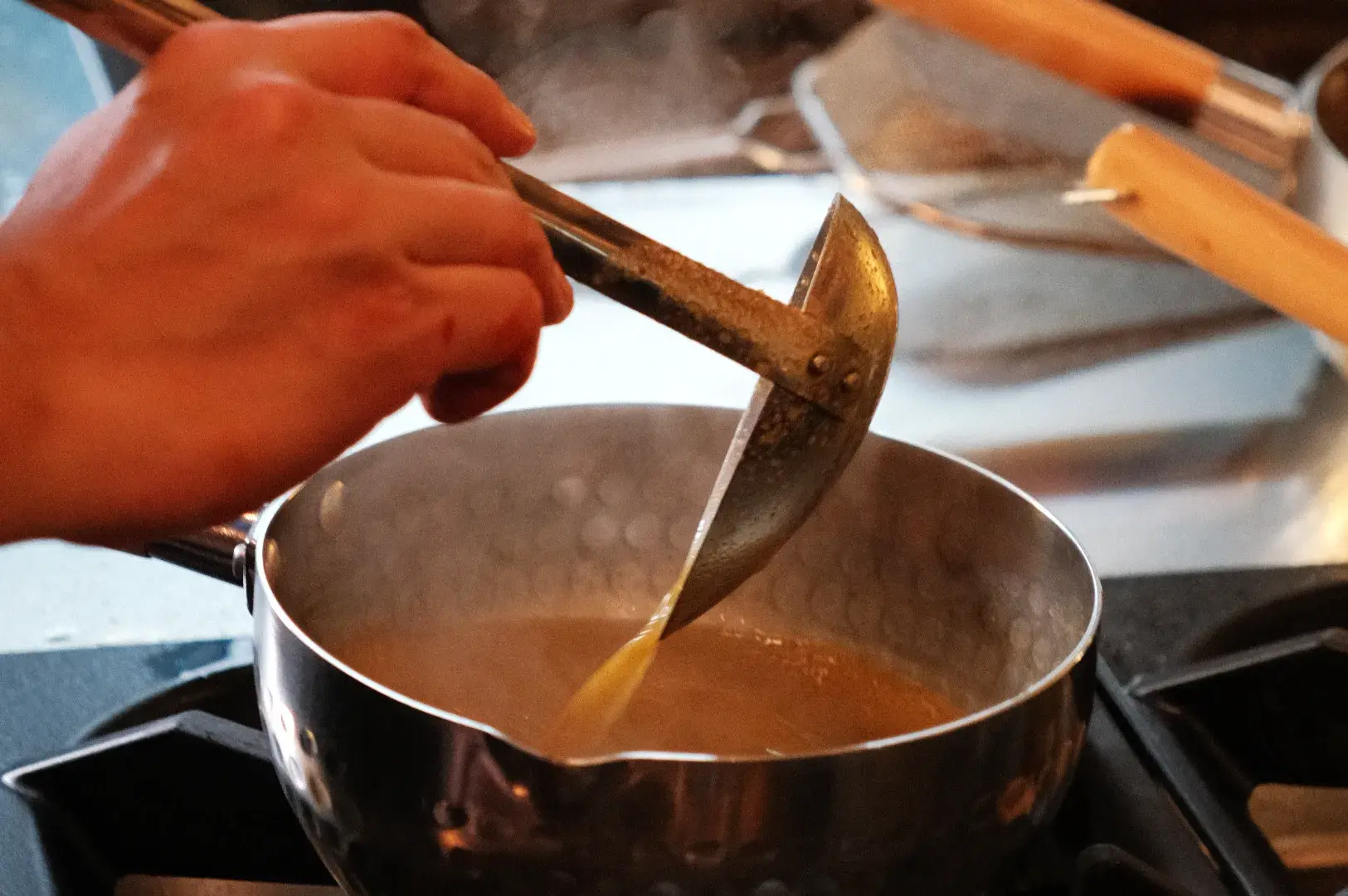 A hand holding a specialized kitchen tool stirring a pot of simmering liquid on a stove.