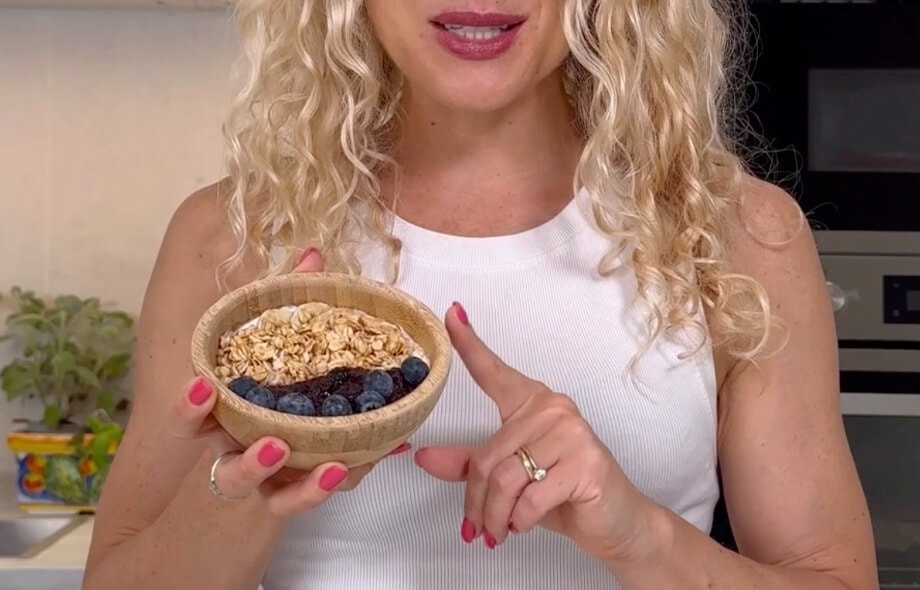 Donna con capelli biondi mossi che tiene nella mano destra una ciotola di muesli con yogurt, mirtilli e marmellata. 