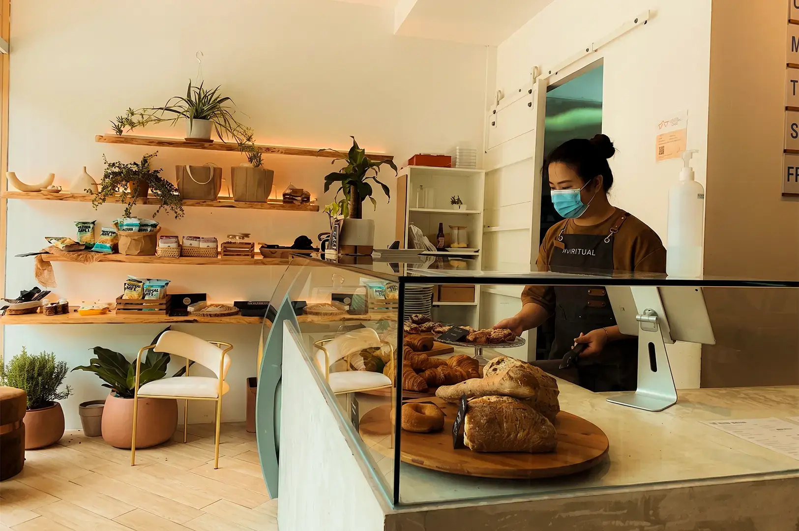 Interior view of The Ritual, with a glass pane of available pastry options displayed in a counter with a staff preparing a meal visible behind. The back wall is decorated with shelved potted plants over a counter and chairs.