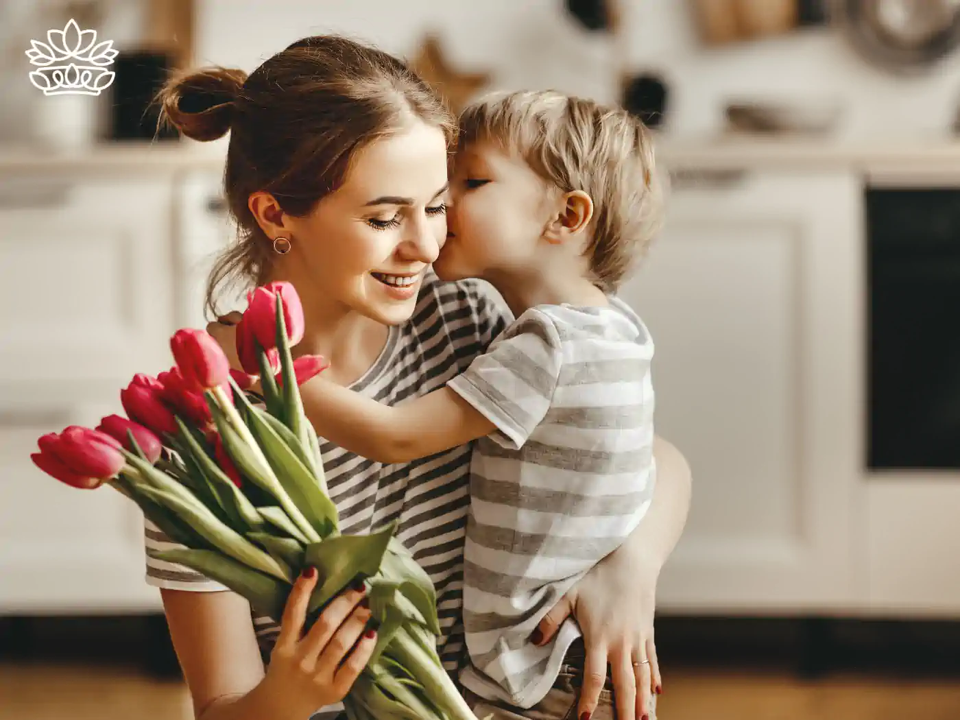 Smiling mother holding her son who kisses her cheek, while she holds a bunch of fresh red tulips in a home setting, showcasing lovingly affordable gifts with flower bouquets under R500 from Fabulous Flowers and Gifts.