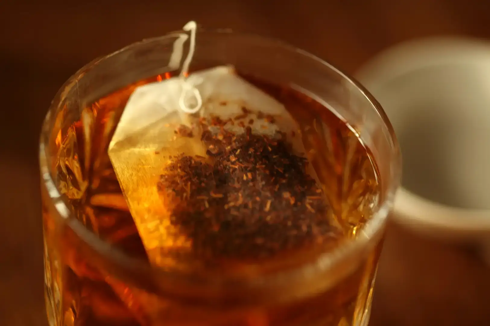 A close-up shot captures a translucent teabag filled with fine, dark rooibos leaves steeping inside a glass filled with golden-orange tea under warm light. The soft-focus background accentuates the rich color and fine texture of the brewing leaves within the glass.