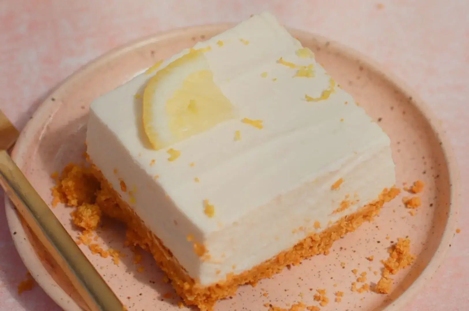 A square slice of lemon dessert on a peach-colored plate with a golden fork beside it.