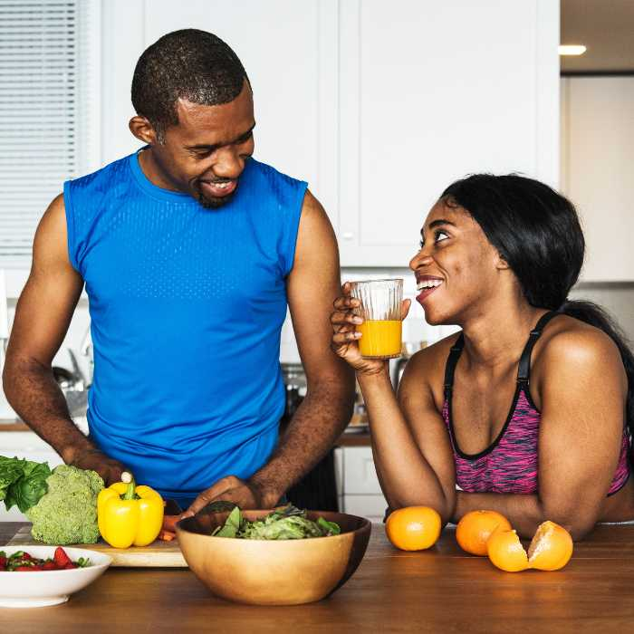A joyful couple engaged in healthy meal preparation in their kitchen, with the man slicing vegetables and the woman enjoying a glass of orange juice. Their vibrant, active lifestyle exemplifies the benefits of AIM Companies Supplements. The Good Stuff Health Shop South Africa.