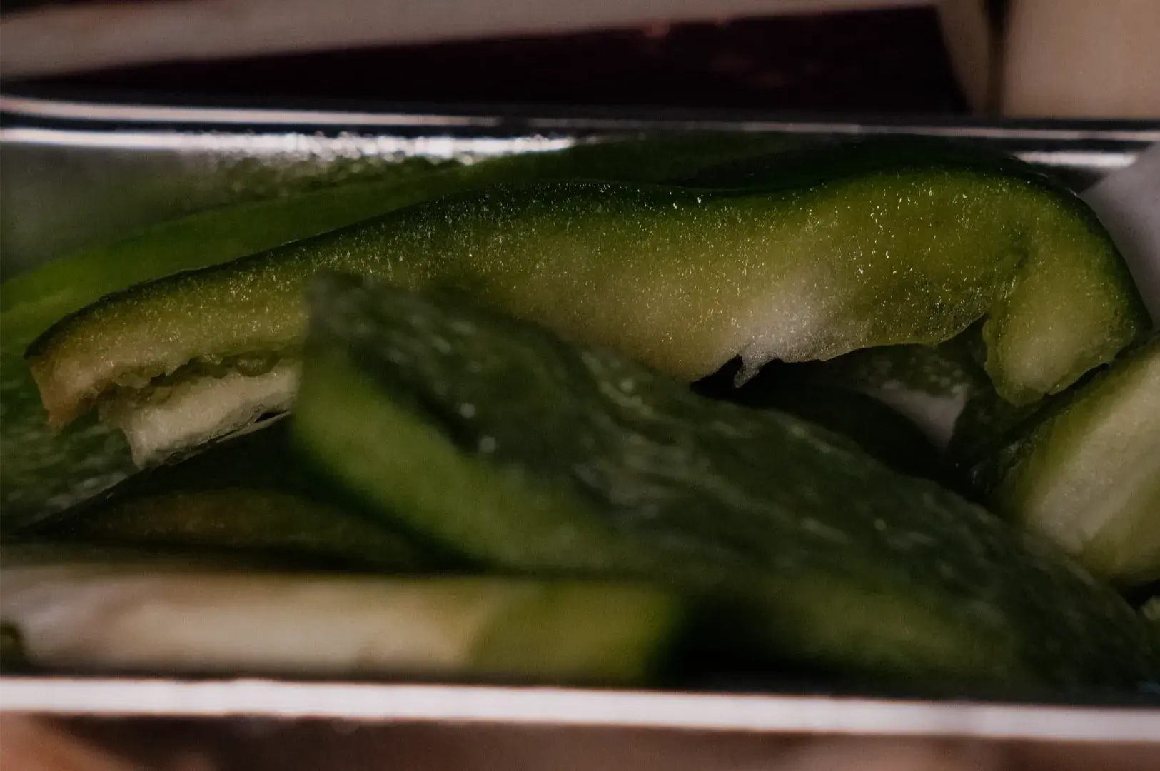 Close-up of glossy green pickled cucumber spears packed in a glass jar, their textured skins catching dim light and creating a moody, savory tone.