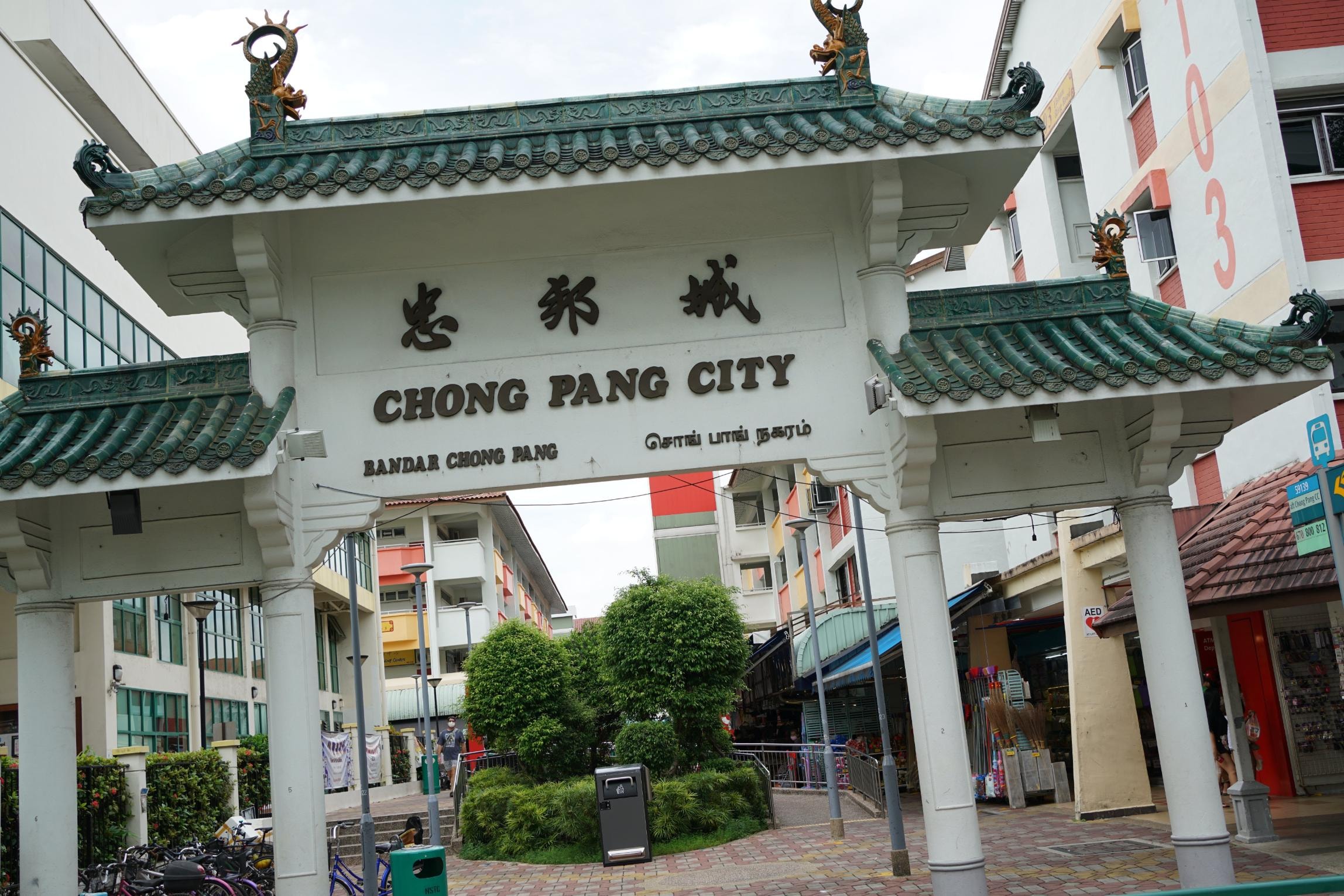  An ornate gateway welcomes visitors to "Chong Pang City," featuring traditional green-tiled roofing adorned with decorative dragon sculptures. The entrance is supported by white pillars and displays the location's name in English, Chinese, Malay, and Tamil above a paved pedestrian area.