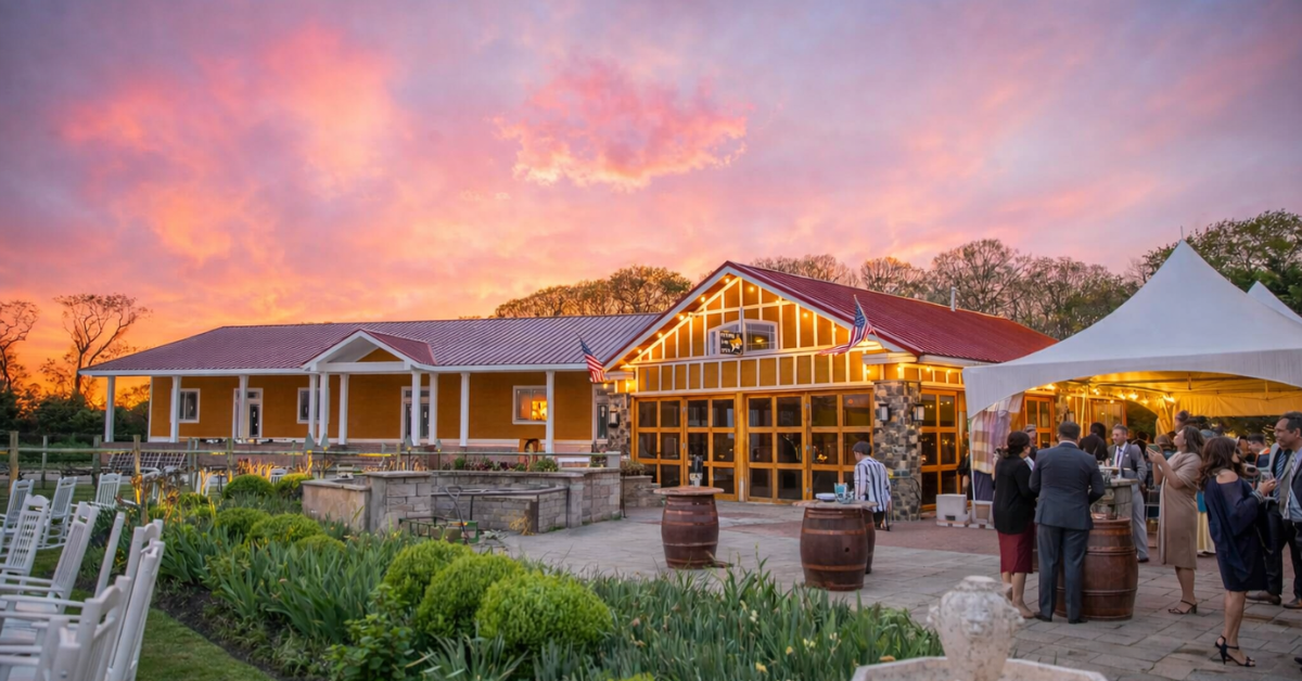 Sunset view of a Cape May vineyard-style event venue with guests gathered outside, warm lighting, landscaped gardens, and a rustic building under a colorful evening sky.