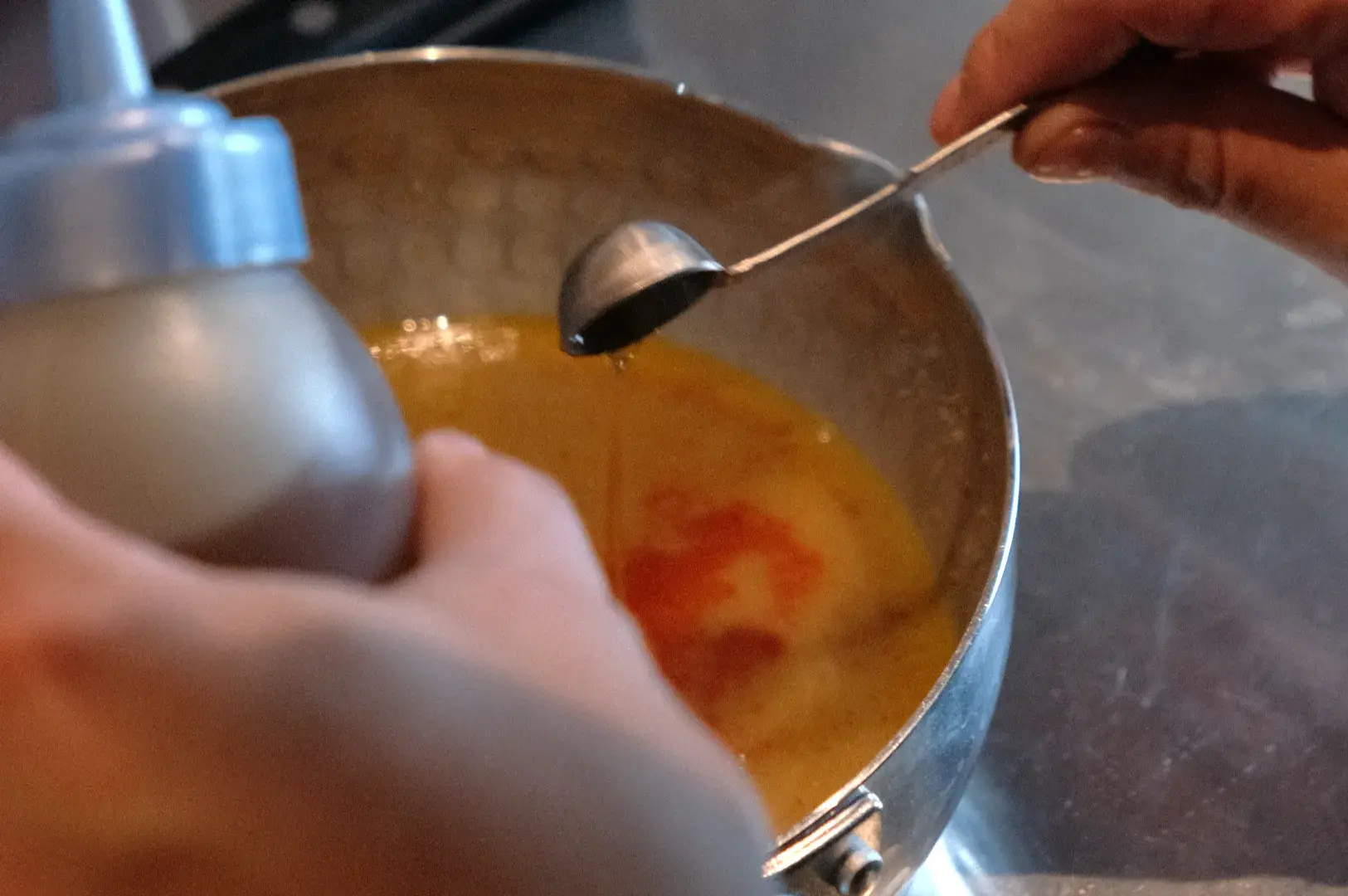 A person holding a spoon and a bottle, pouring a liquid into a metal bowl containing a yellow mixture.