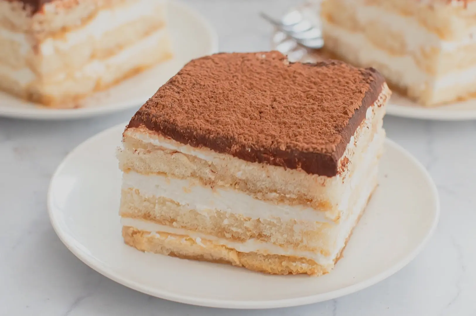A slice of layered dessert topped with cocoa powder, sitting on a white plate. Other similar dessert slices are in the background.