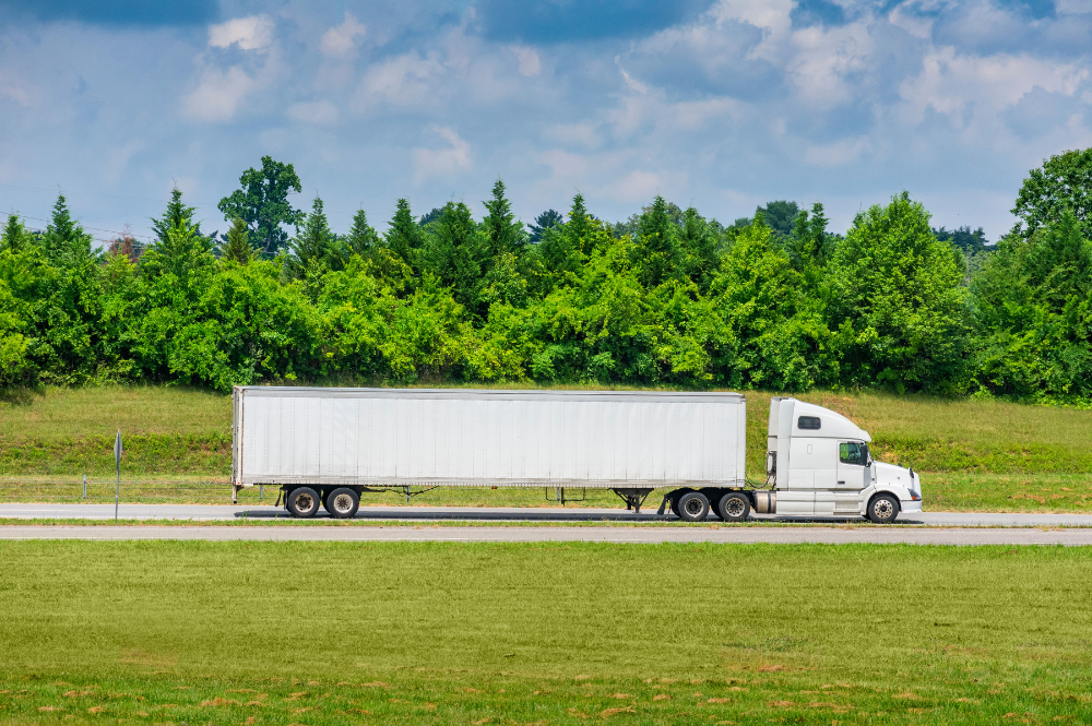 A semi truck travels along a road beside a lush green field, illustrating transportation equipment in use.