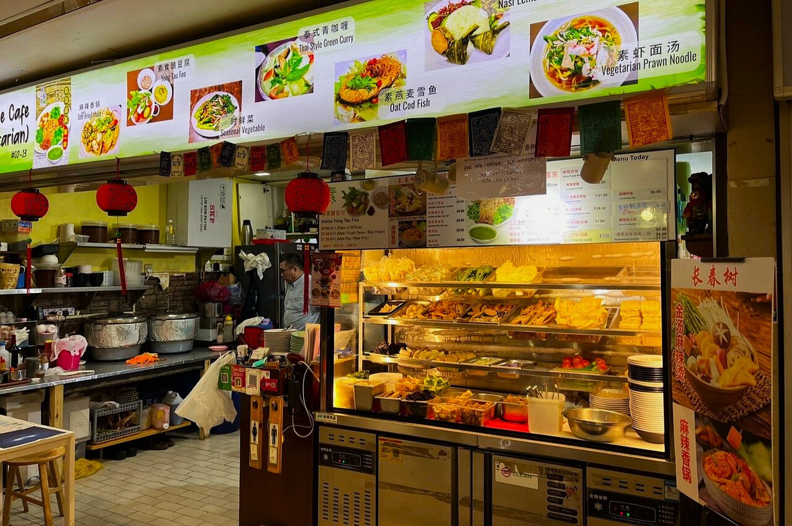 A bustling vegetarian food stall with colorful light-up menu displaying dishes like Thai Green Curry and Vegetarian Prawn Noodle. Warm, inviting atmosphere.
