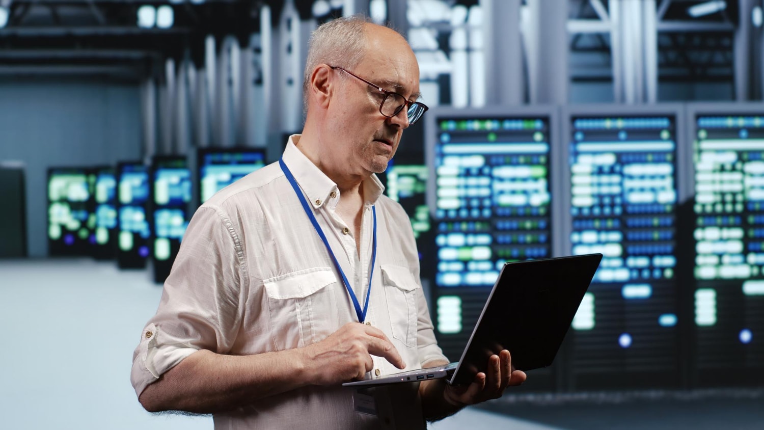 A man in a data center uses a laptop to oversee telecom security and business phone systems. The illuminated server racks suggest cloud-based telecom and call routing solutions.