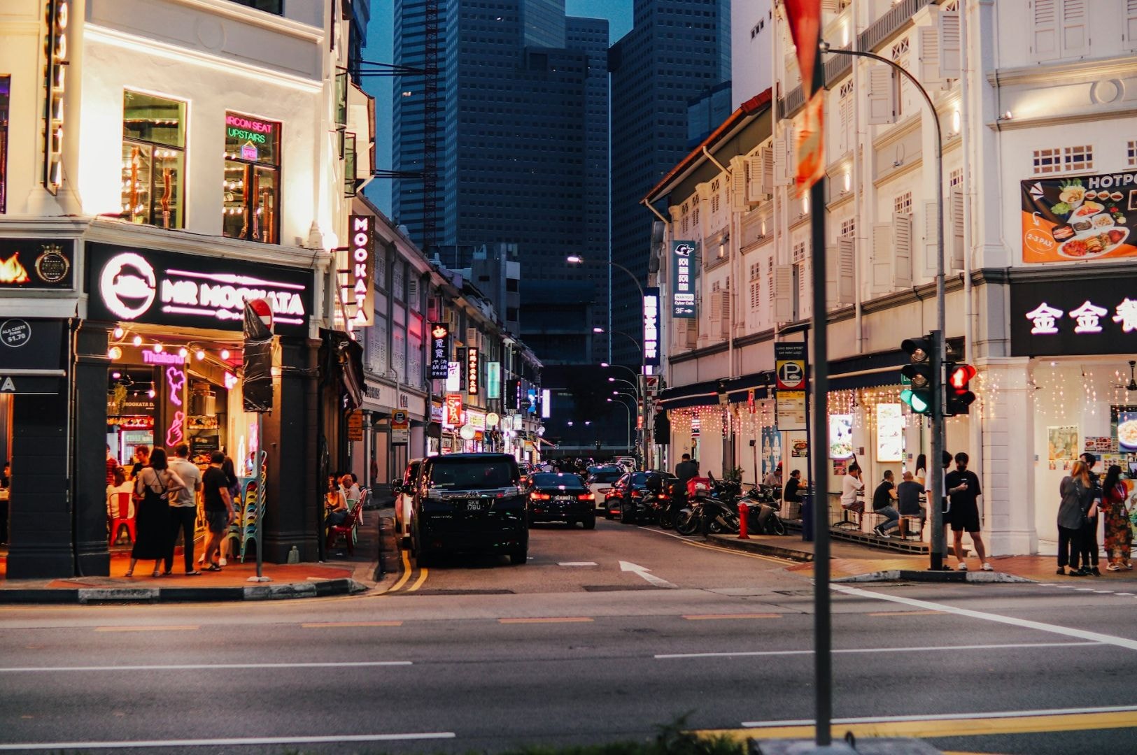 A bustling city street at dusk, lined with brightly lit shops and restaurants. People walk on sidewalks, and skyscrapers loom in the background, exuding a vibrant urban energy.