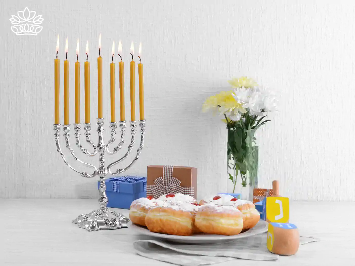 A lit menorah with nine yellow candles, placed on a table alongside wrapped gifts, traditional Hanukkah doughnuts, and a vase with white and yellow flowers. Fabulous Flowers and Gifts - Hanukkah Flowers.