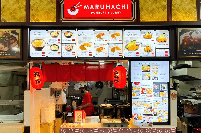 Front view of the Maruhachi Donburi & Curry food stall, featuring brightly illuminated menu boards displaying various Japanese curry dishes, traditional red noren curtains, hanging red paper lanterns, and a staff member working behind the counter.