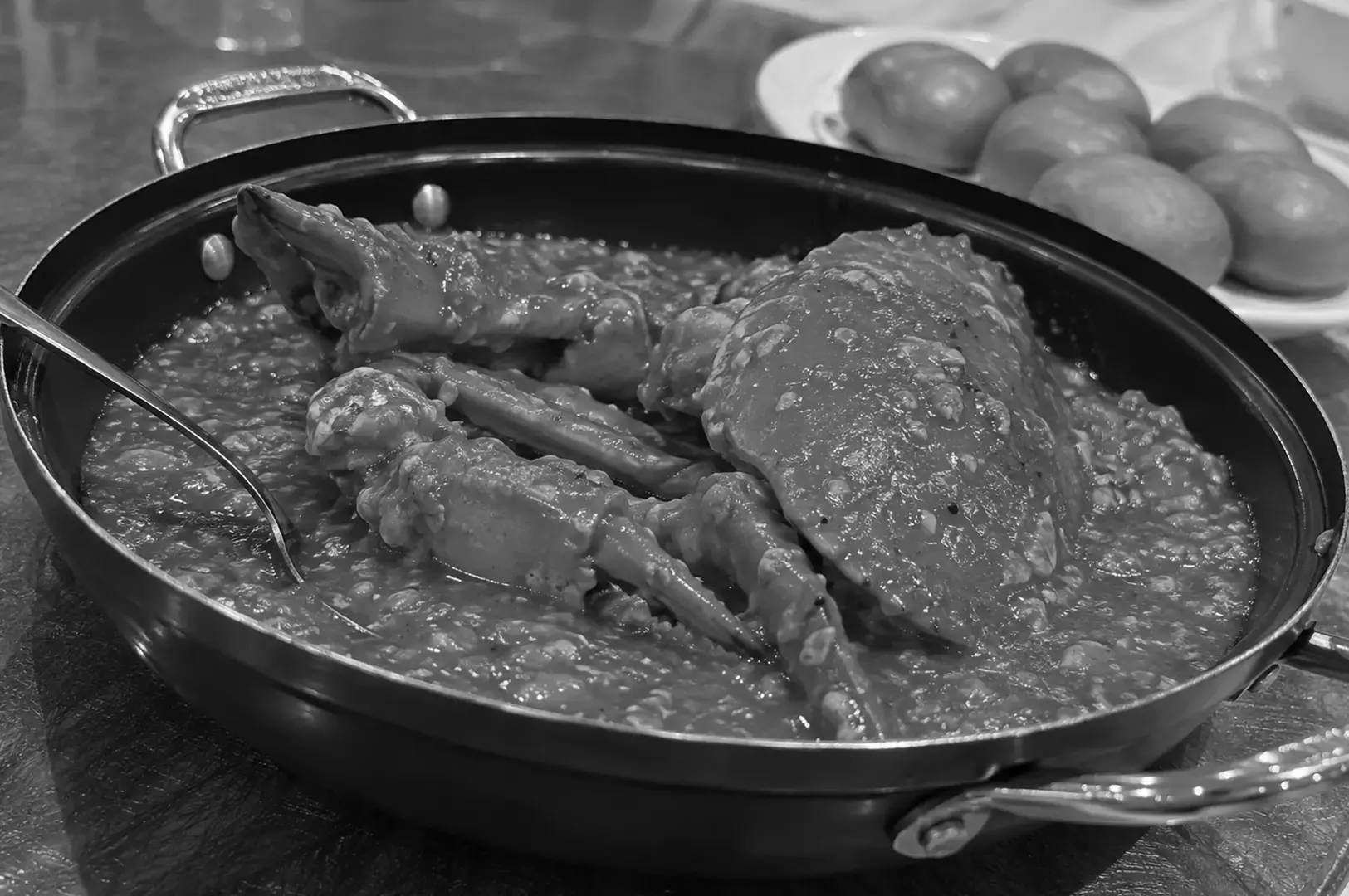 Whole crab cooked in a thick, textured sauce inside a black round serving pan, with a metal serving spoon resting along the side and a plate of buns in the background.
