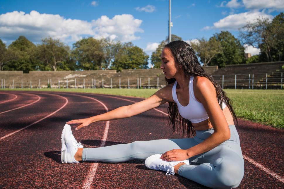 Runner sitting on a track field doing a seated hamstring stretch, reaching toward the toes with one leg extended.