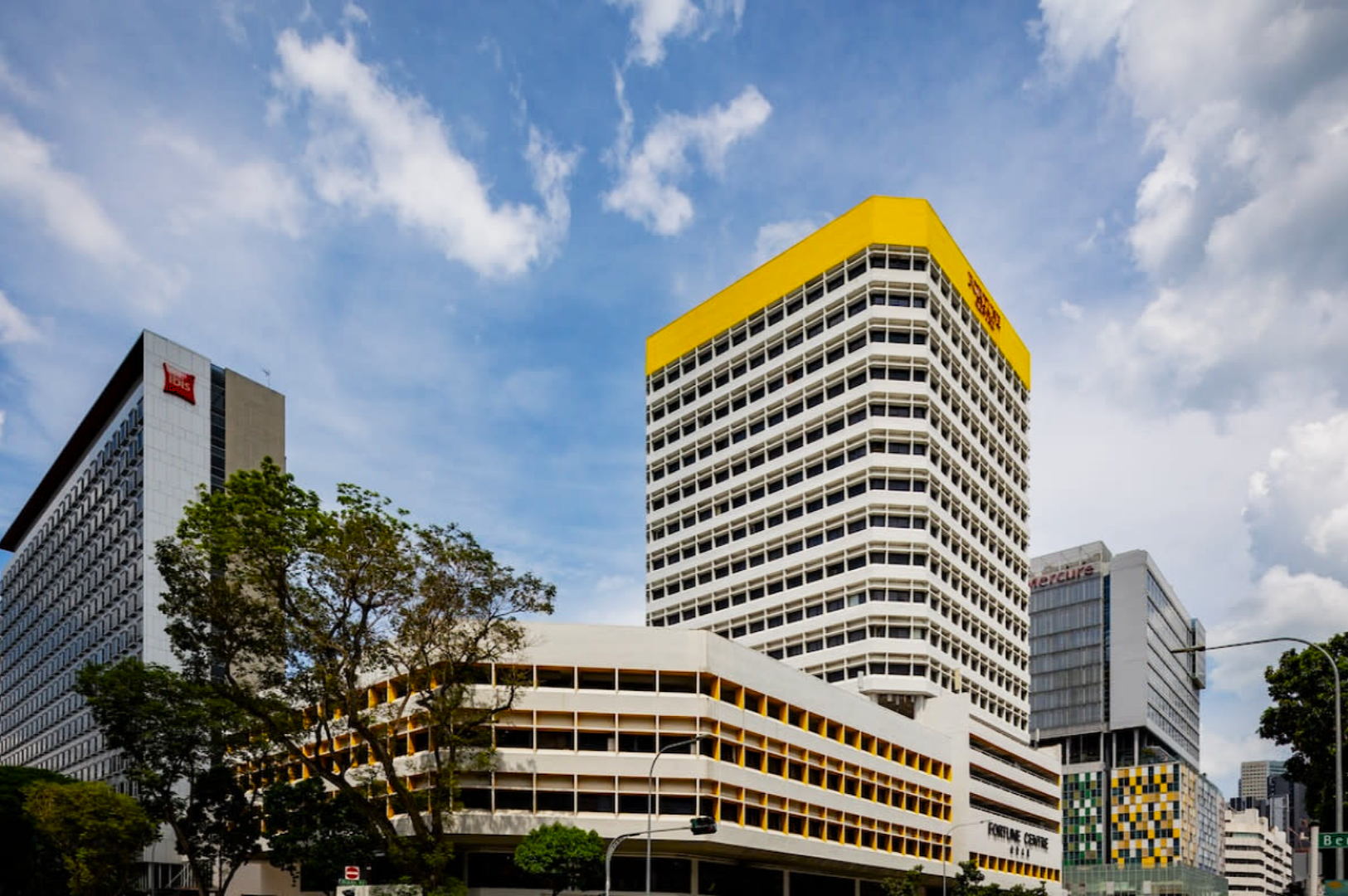 Cityscape with modern buildings under a partly cloudy sky. The tallest building features white grid-like windows and a bright yellow top, conveying a vibrant urban feel.