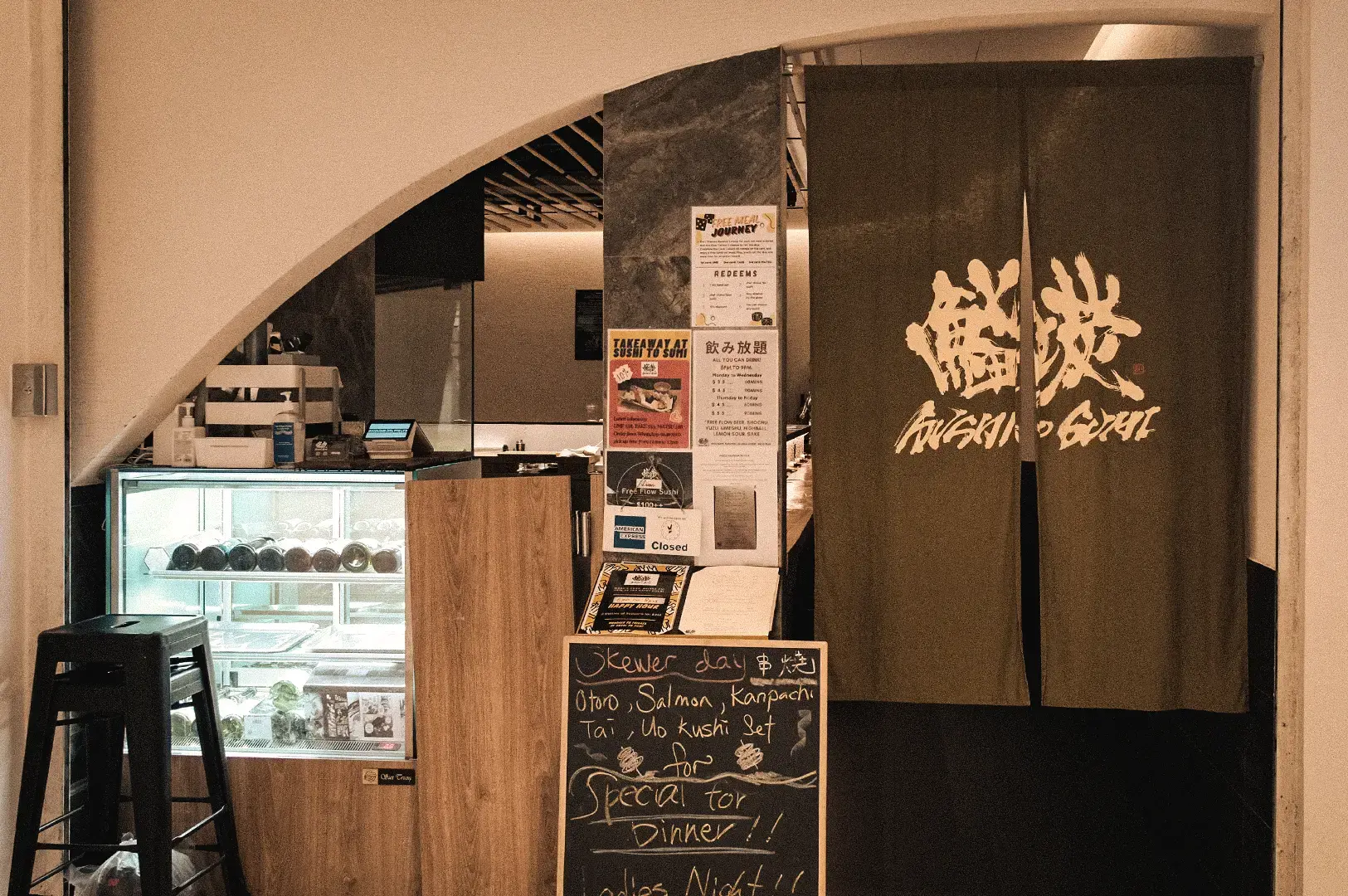 An eye-level medium shot reveals the entrance to Sushi to Sumi, a standing sushi bar in Raffles Place featuring a dark noren curtain and handwritten daily menu board.