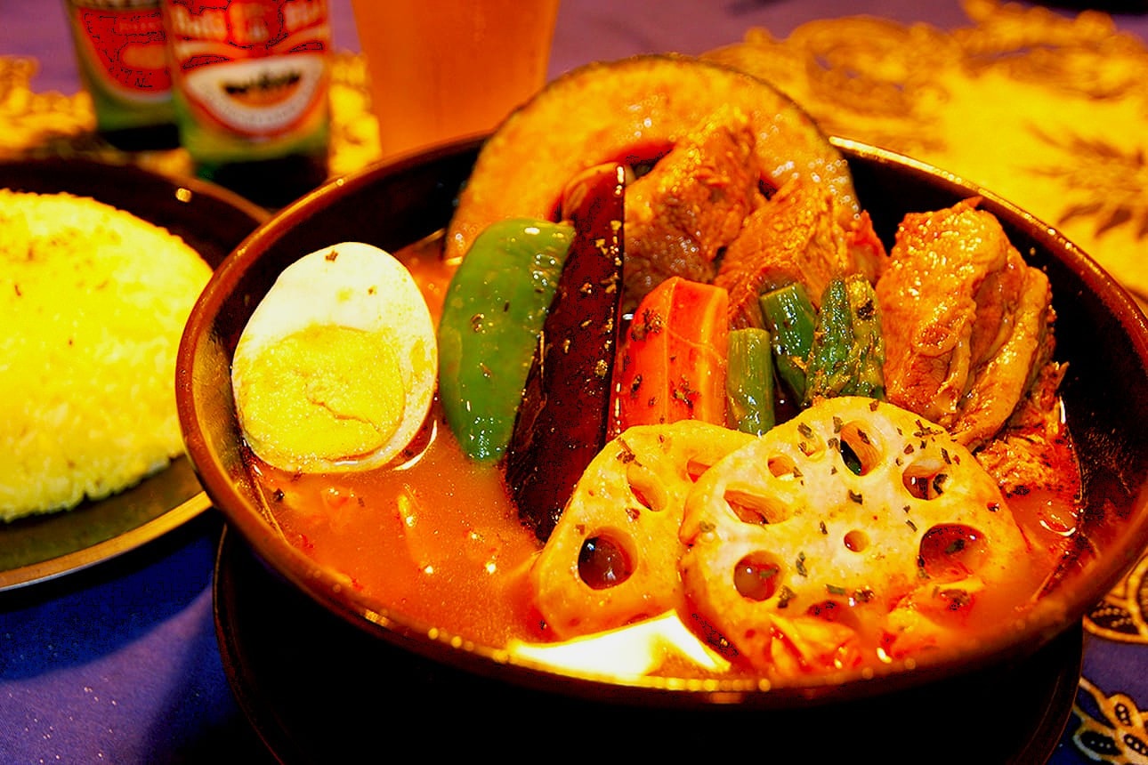 A vibrant Japanese curry dish featuring boiled egg, lotus root, peppers, carrots, and chicken in a rich broth, served with yellow rice.