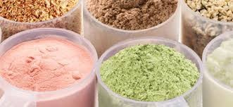 Several scoops of various protein powders, including pink, green, brown, and beige.