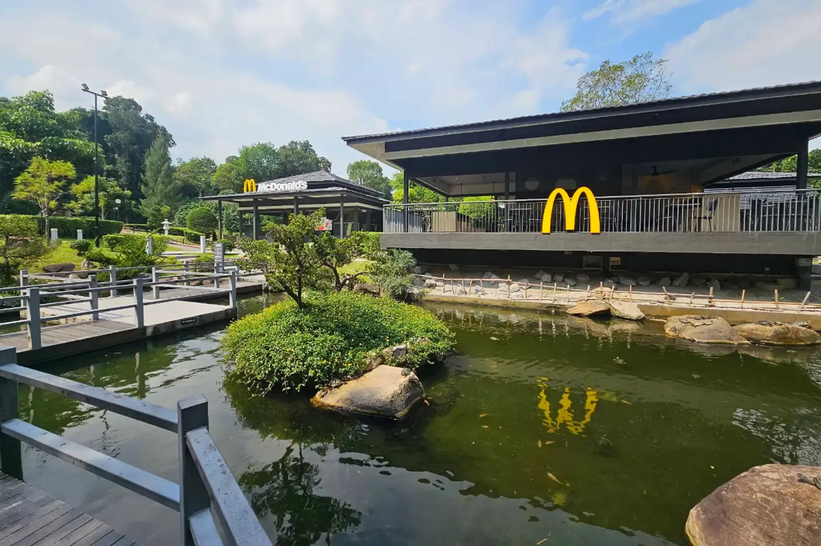 A wide daytime view of a dark-timber, Japanese-style McDonald's pavilion featuring the iconic golden arches along its balcony railing, situated beside a calm green pond with a small landscaped island, wooden footbridges, and lush surrounding foliage under a bright sky.