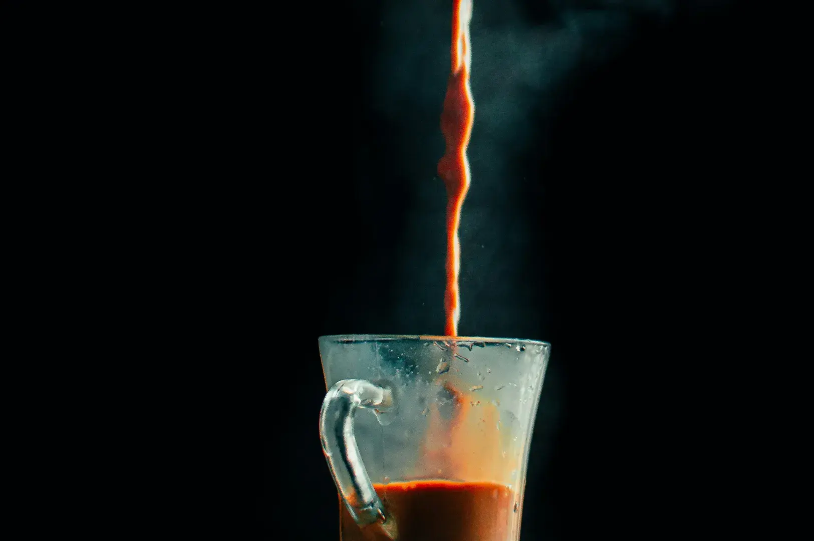 Stream of hot milk tea pouring into a glass mug against a black background.