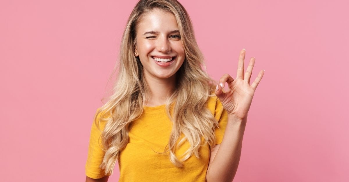 Blonde woman in a yellow shirt smiling and winking, giving an "OK" hand sign – tax deductions list for self employed.