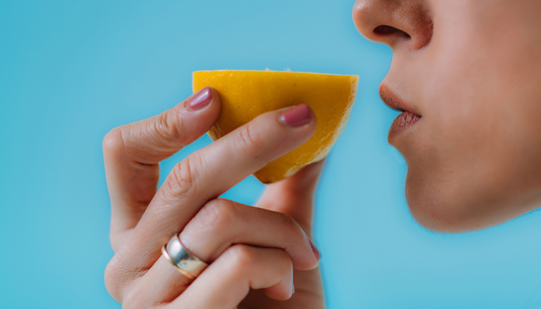 Person holder en halv sitron nær munnen, klar til å smake eller lukte på den. Blå bakgrunn gir kontrast til den gule frukten.