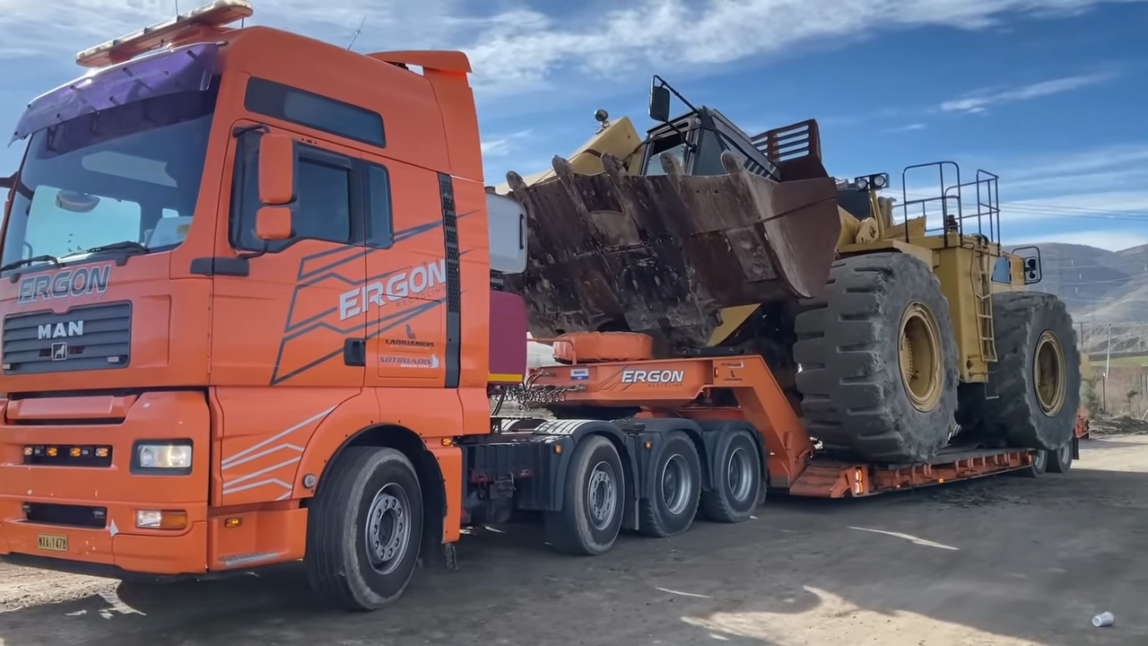 An orange ERGON branded heavy-duty truck carrying an oversized yellow construction vehicle with large wheels and a rusty scoop on the back of the trailer