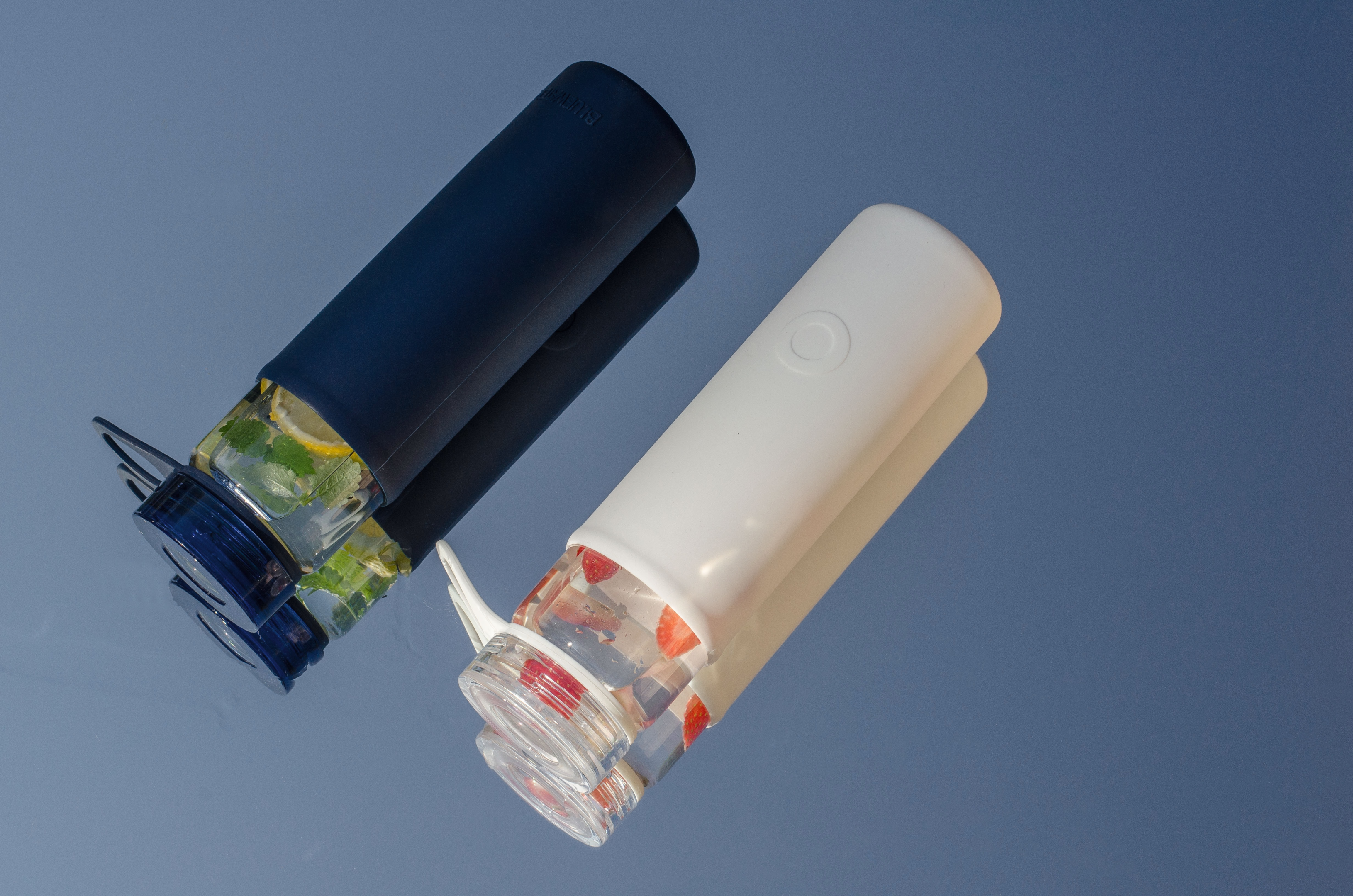 Deux gourdes en plastique sans bpa. L'une est noire, l'autre est blanche. Les deux contiennent de l'eau aromatisé au citron (pour la gourde noire) et de fraise (pour la gourde blanche). Les deux sont sur un fond bleu dégradé.